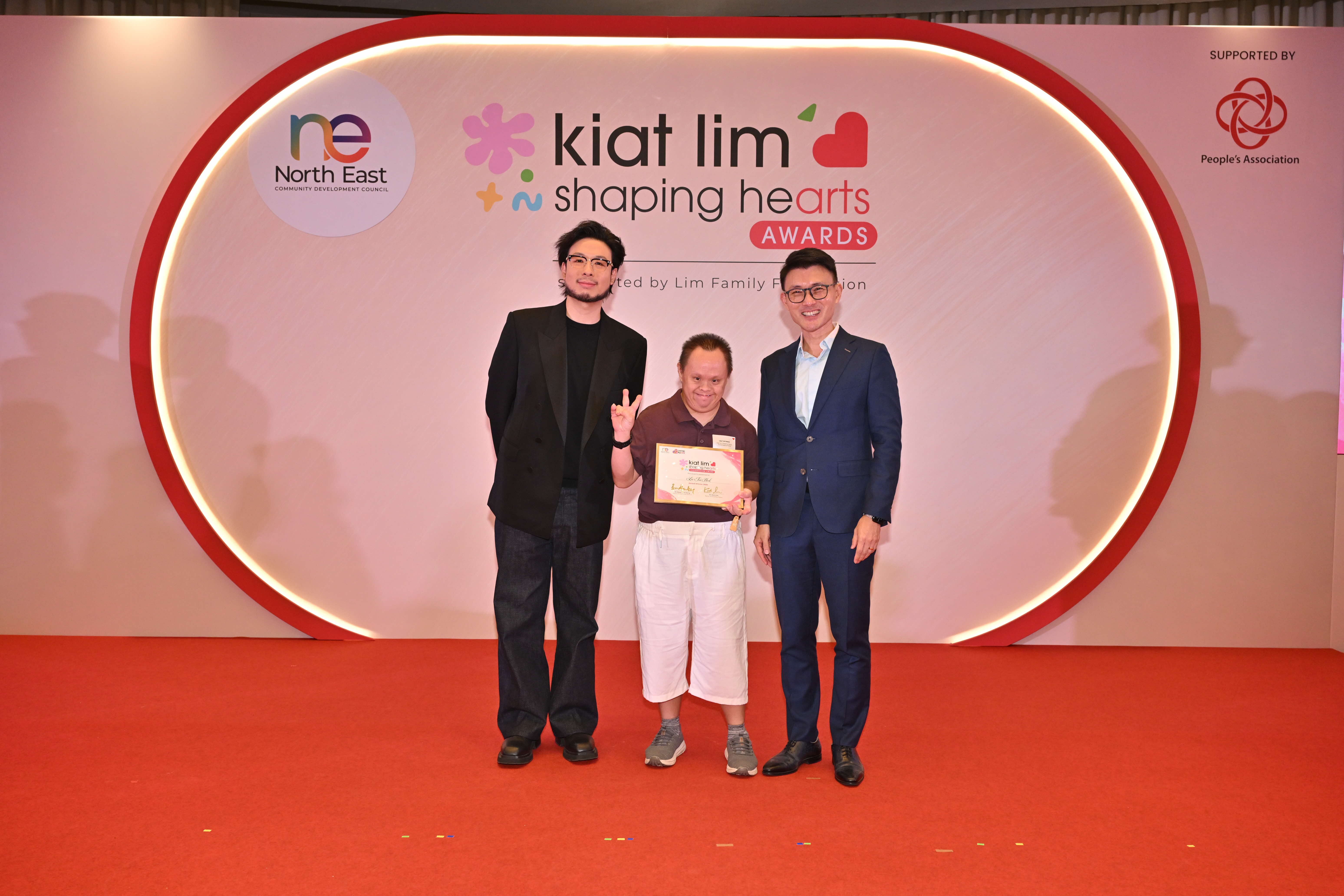 Award recipient standing on stage with the presenter, holding their certificate and posing for a group photo during the award ceremony, with a backdrop reading ‘Kiat Lim-Shaping Hearts Awards'.